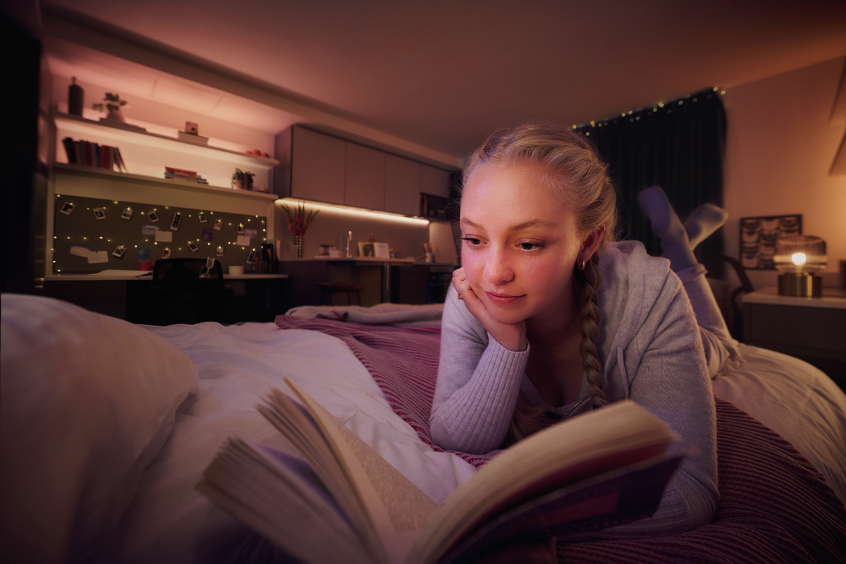 A person with braided hair is lying on a bed, reading a book. The room is cozy with dim lighting, and there are shelves with books and decorative lights in the background.